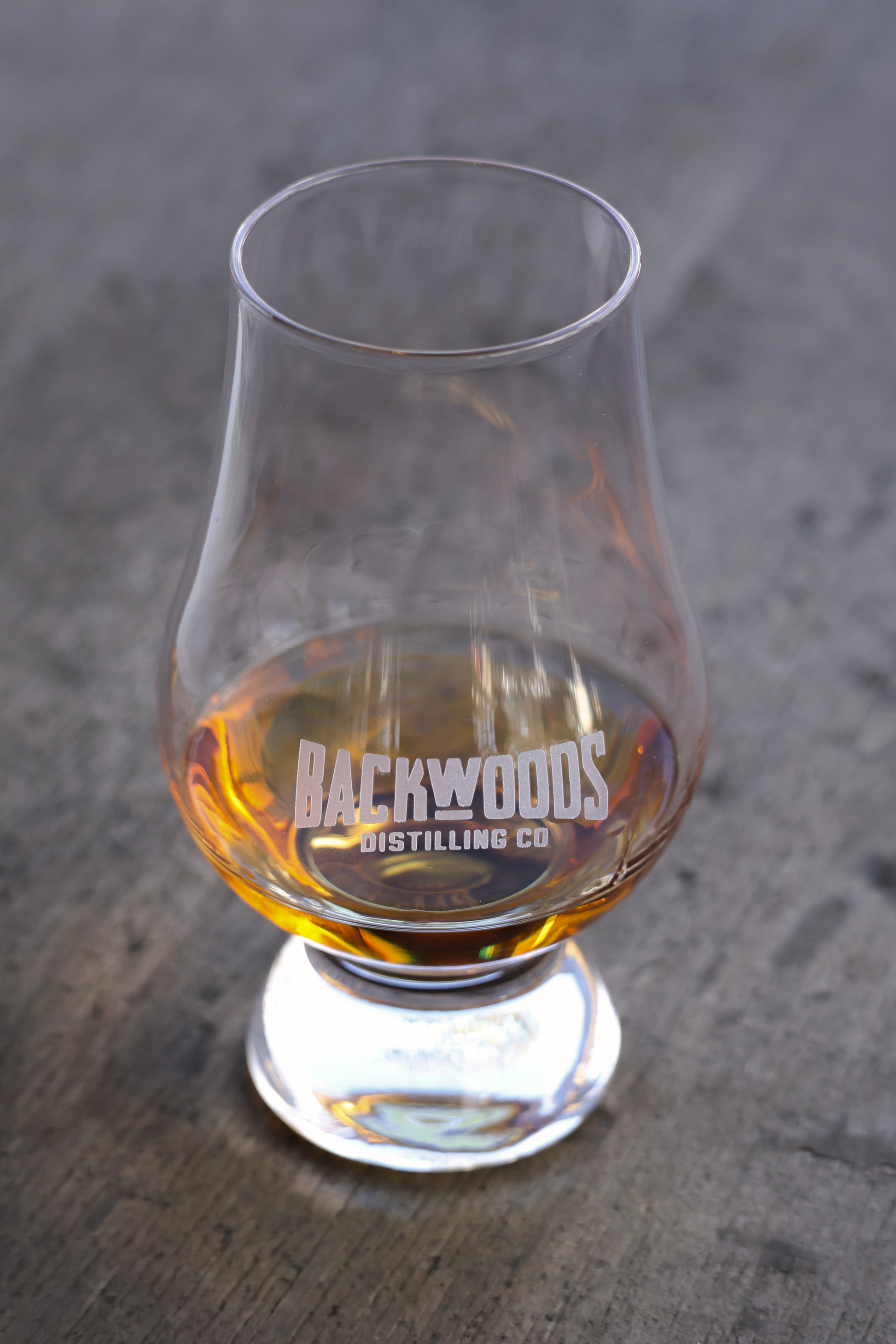A clear whisky glass with the brand name 'BACKWOODS DISTILLING CO' printed on it, filled with amber liquor, set on a gray wooden surface.
