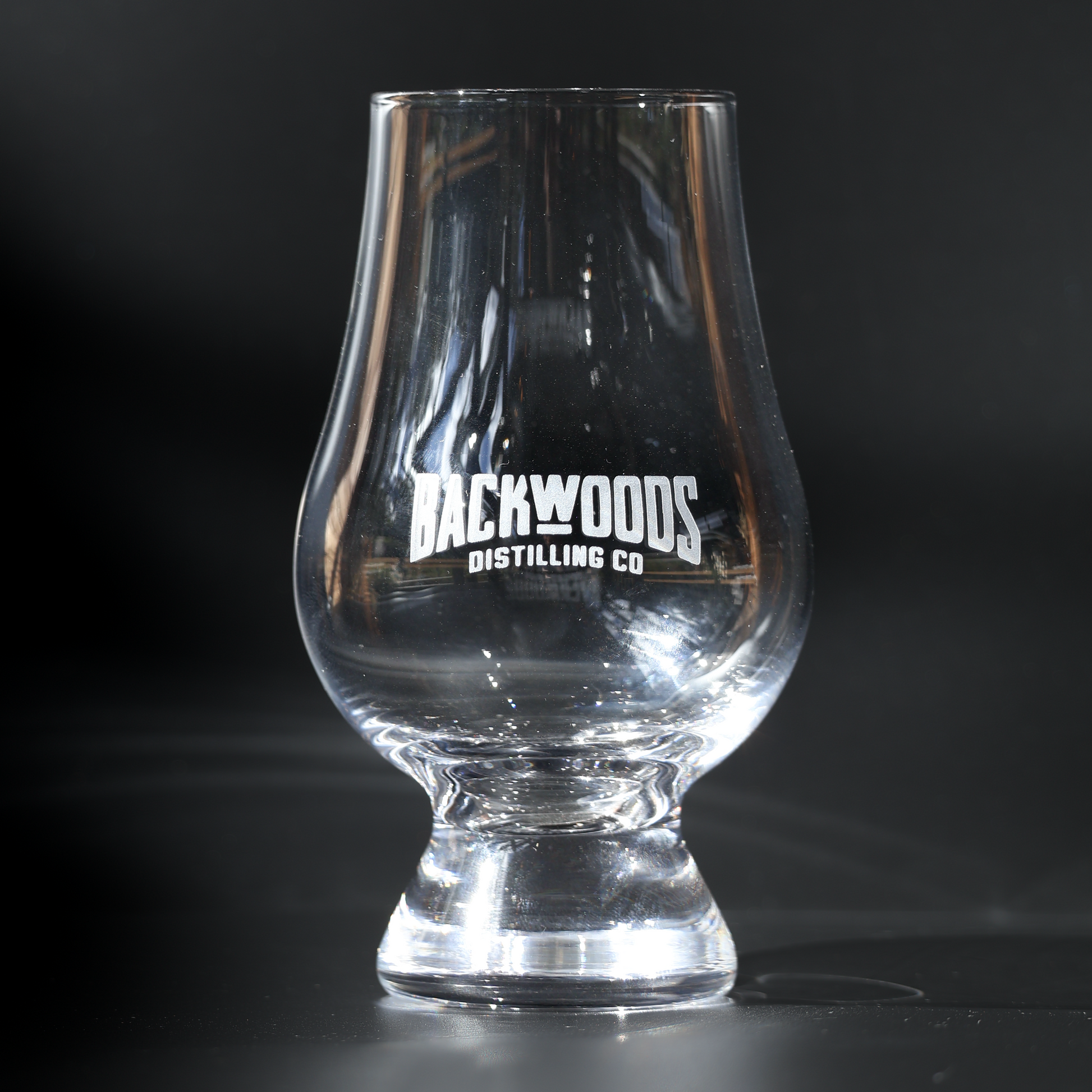 A clear glass tulip-shaped tumbler with the white text 'BACKWOODS DISTILLING CO' printed on the front, set against a dark, reflective background.