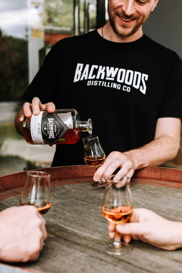 Person in a Backwoods shirt pouring whisky from a Backwoods bottle into a tasting glass.