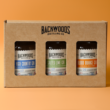 A cardboard gift box containing three Backwoods Distilling Co. gin bottles, shown against a solid orange background.
