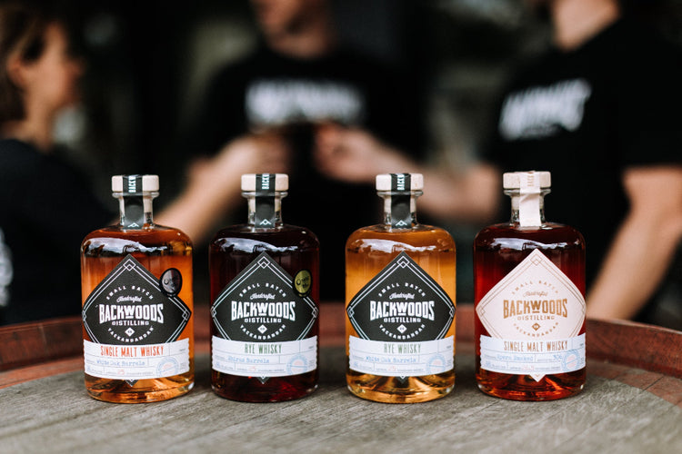 Four bottles of Backwoods whisky on a wooden surface with people in the background.