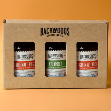 A three-pack of Backwoods Distilling Co. single malt and rye whiskies in a brown cardboard gift box with three clear windows showing the bottles inside, set against a solid orange background.