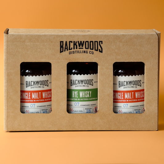 A three-pack of Backwoods Distilling Co. single malt and rye whiskies in a brown cardboard gift box with three clear windows showing the bottles inside, set against a solid orange background.