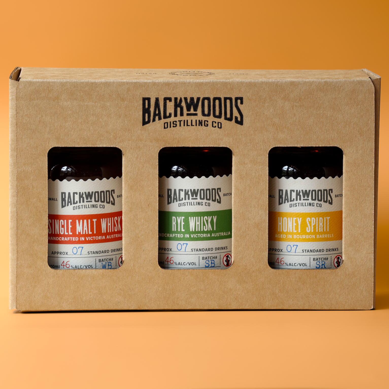 A three-pack of Backwoods Distilling Co. whiskies in a brown cardboard gift box, set against a solid orange background.