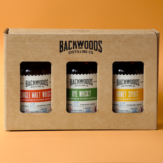 A three-pack of Backwoods Distilling Co. whiskies in a brown cardboard gift box, set against a solid orange background.